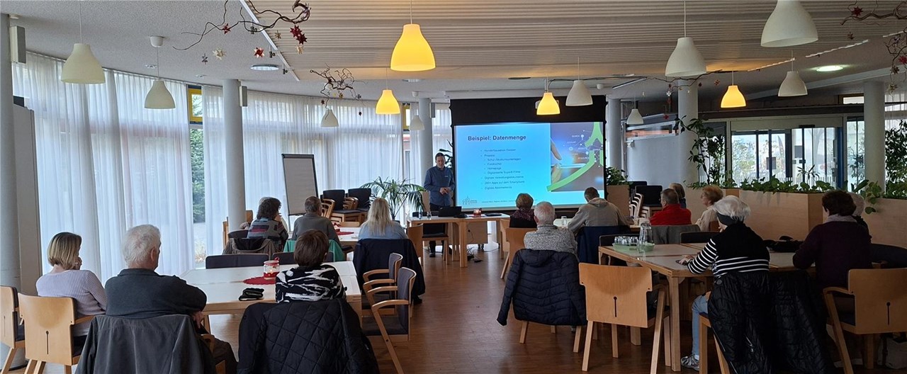 Das Bild zeigt Johannes Baur an einer Leinwand stehend, auf der eine Präsentation zu sehen ist. Im Vordergrund vier Tische mit Gästen der Veranstaltung