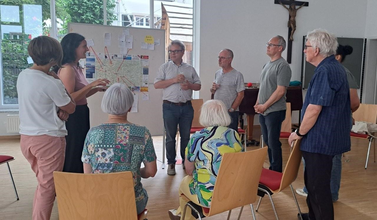 Eine Gruppe von Menschen stehen und sitzen im Kreis, sie schauen sich an und unterhalten sich miteinander. Im Hintergrund ist eine Stellwand mit einer Landkarte, Post-its und Pfeilen zu sehen.