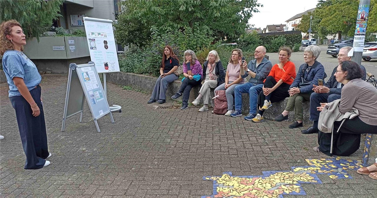 Eine Person steht neben zwei Stellwänden und erläutert etwas, ca. 10 Teilnehmende sitzen im Halbkreis und hören zu.