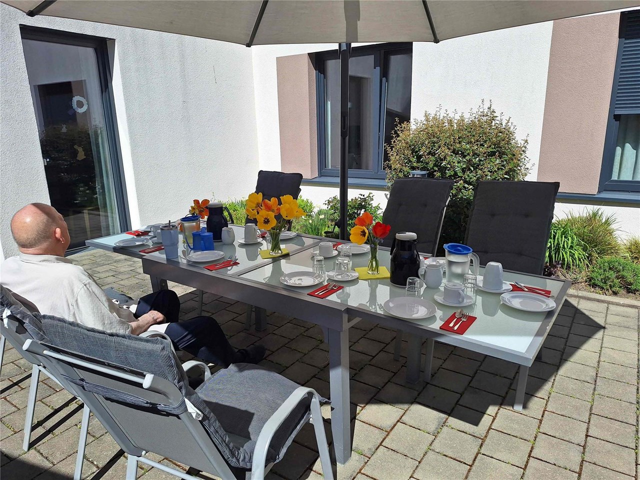 Ein gedeckter Tisch mit Blumen und Geschirr steht auf einer sonnigen Terrasse unter einem Sonnenschirm. Mehrere Stühle sind frei, eine Person sitzt am Tisch.
