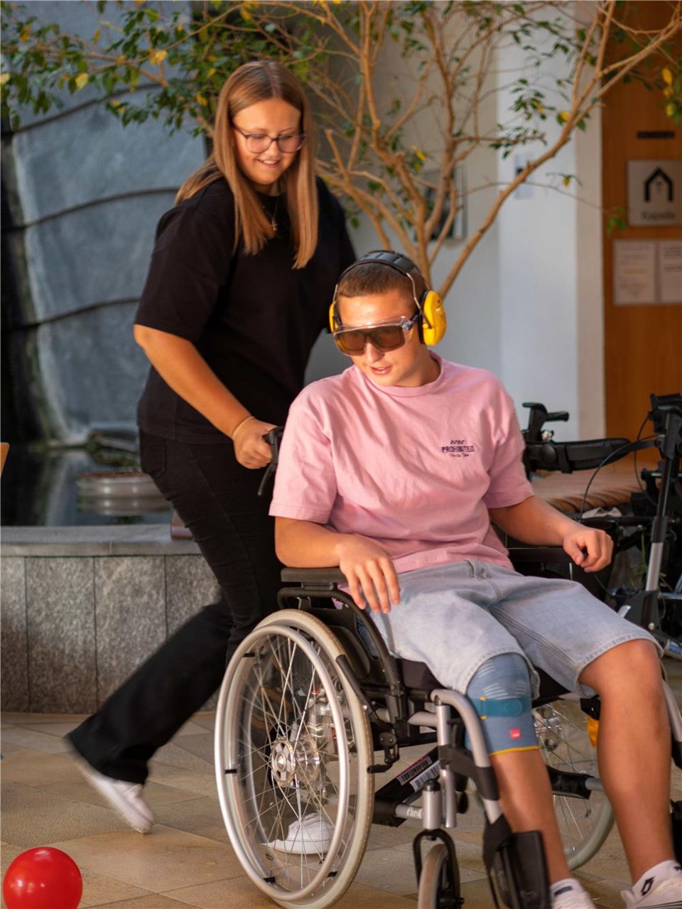 Auf dem Bild sind zwei junge Menschen zu sehen, einer davon sitzt im Rollstuhl mit verdunkelter Brille und Kopfhörer, die andere Person schiebt den Rollstuhl.