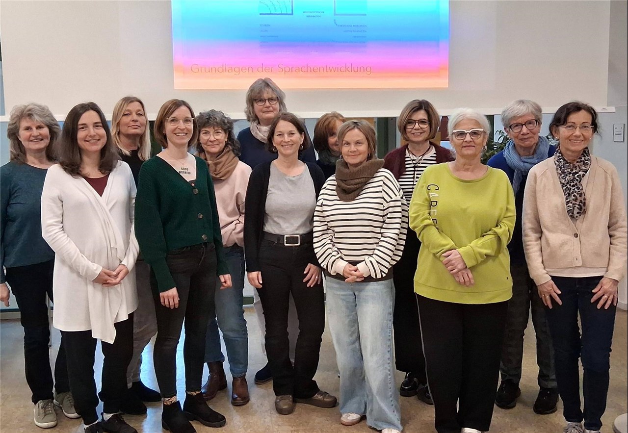 13 Mitarbeiter*innen der Schulkindbetreuung stehen zusammen, im Hintergrund an einer Leinwand ist eine Folie einer Präsentation mit dem Titel „Grundlagen der Sprachentwicklung“ zu sehen