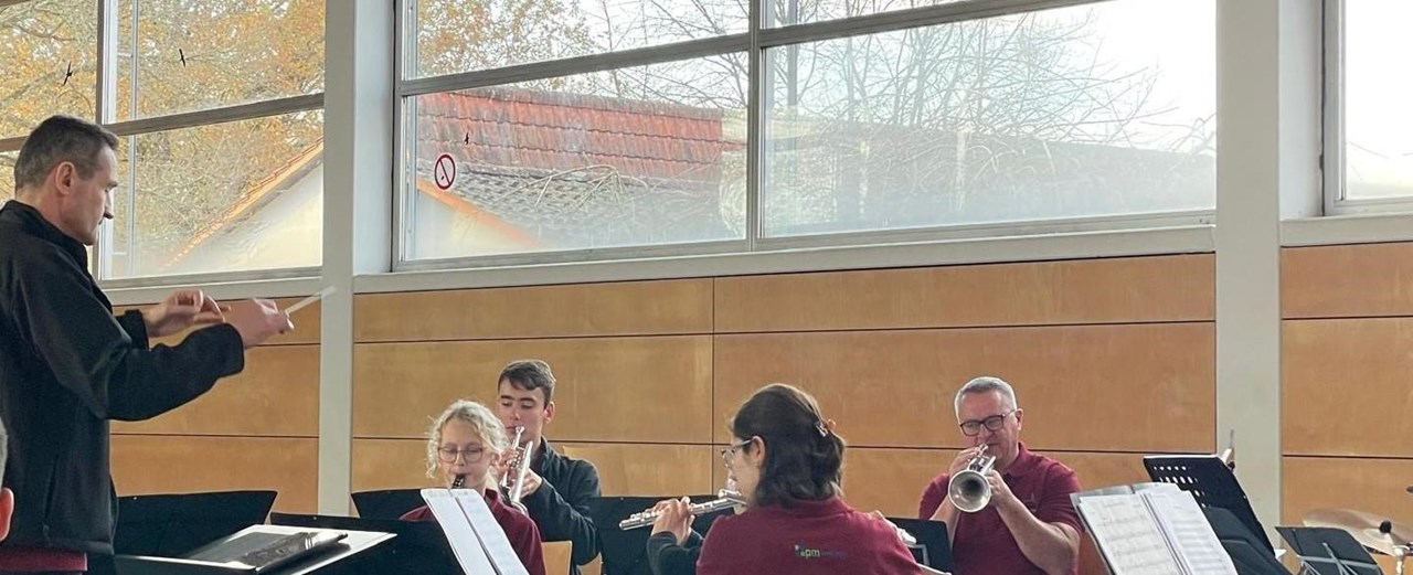 Das Bild zeigt am unteren Rand vier Musizierende mit Blasinstrument. Rechts ist der Dirigent zu sehen. Oberhalb ist ein großes Fenster, man sieht ein Dach und Bäume