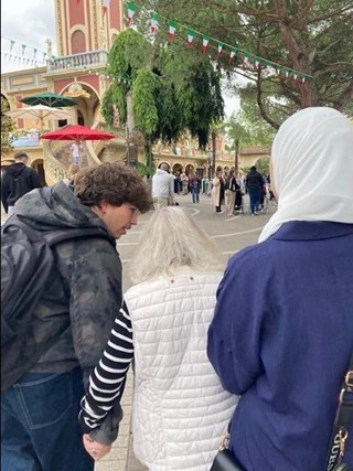 Drei Personen, davon eine ältere Dame, sind seitlich oder von hinten zu sehen. Im Hintergrund sieht man einen Teil einer Kulisse des Europaparks.