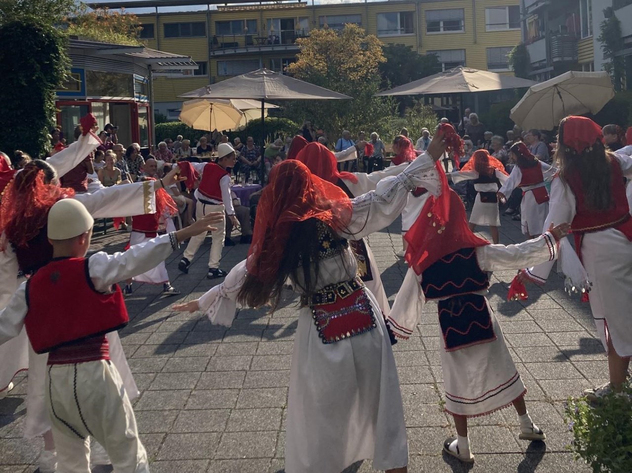 Auf dem Bild ist der Innenhof des Seniorenzentrums Bürkle-Bleiche zu sehen, im Vordergrund tanzen Kinder einer Tanzgruppe in kosovarischer Tracht, im Hintergrund sitzen die Senior*innen und Gäste als Publikum.