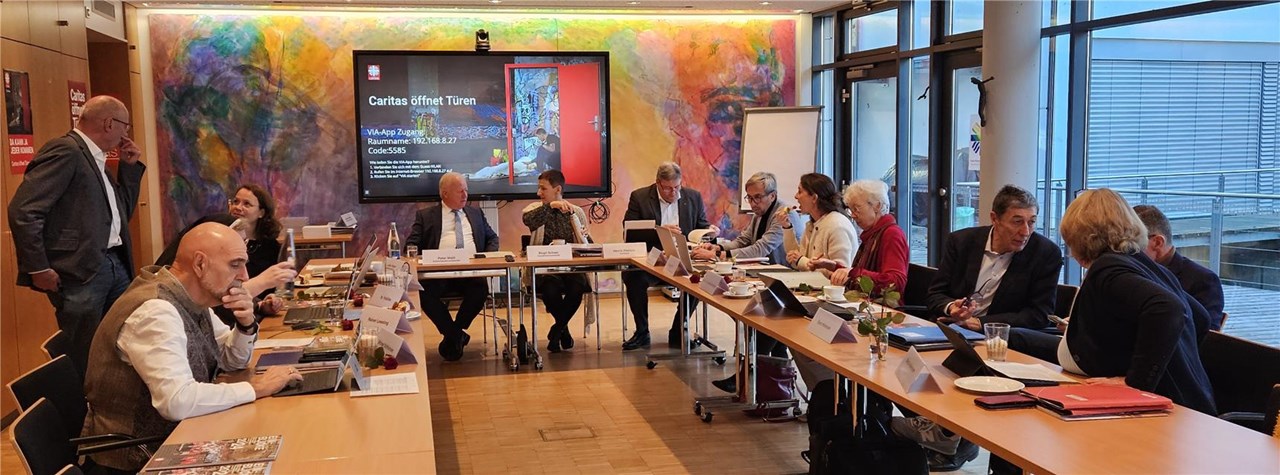 Das Bild zeigt die Mitglieder des Aufsichtsrates und den Vorstand des Diözesancaritasverbandes  (ca. 15 Personen) an Tischen sitzen, die in U-Form gestellt sind. Die Wand im Hintergrund ist bunt, davor ein Bildschirm mit dem Jahresmotto der Caritas „Caritas öffnet Türen“