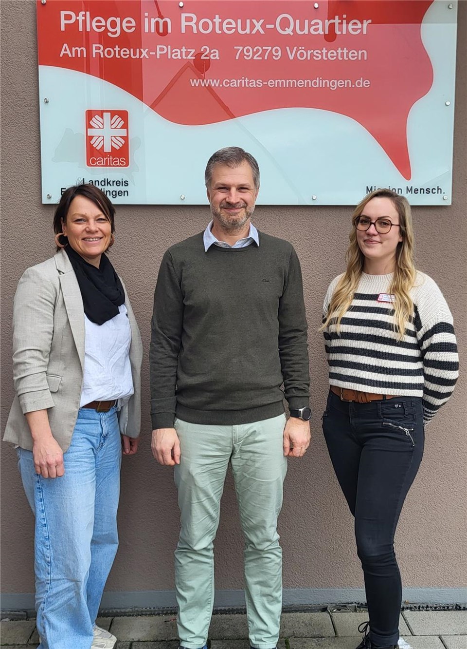 Drei erwachsene Personen stehen lächelnd nebeneinander vor einer Hauswand im Freien. Von links nach rechts: Katja Rambach, Bereichsleitung Altenhilfe, Kevin Schüller, Bürgermeister von Vörstetten und Ines Hanselmann, Pflegefachliche Leitung. Hinter ihnen hängt ein rot-weißes Schild der Caritas mit der Aufschrift „Pflege im Roteux-Quartier“.