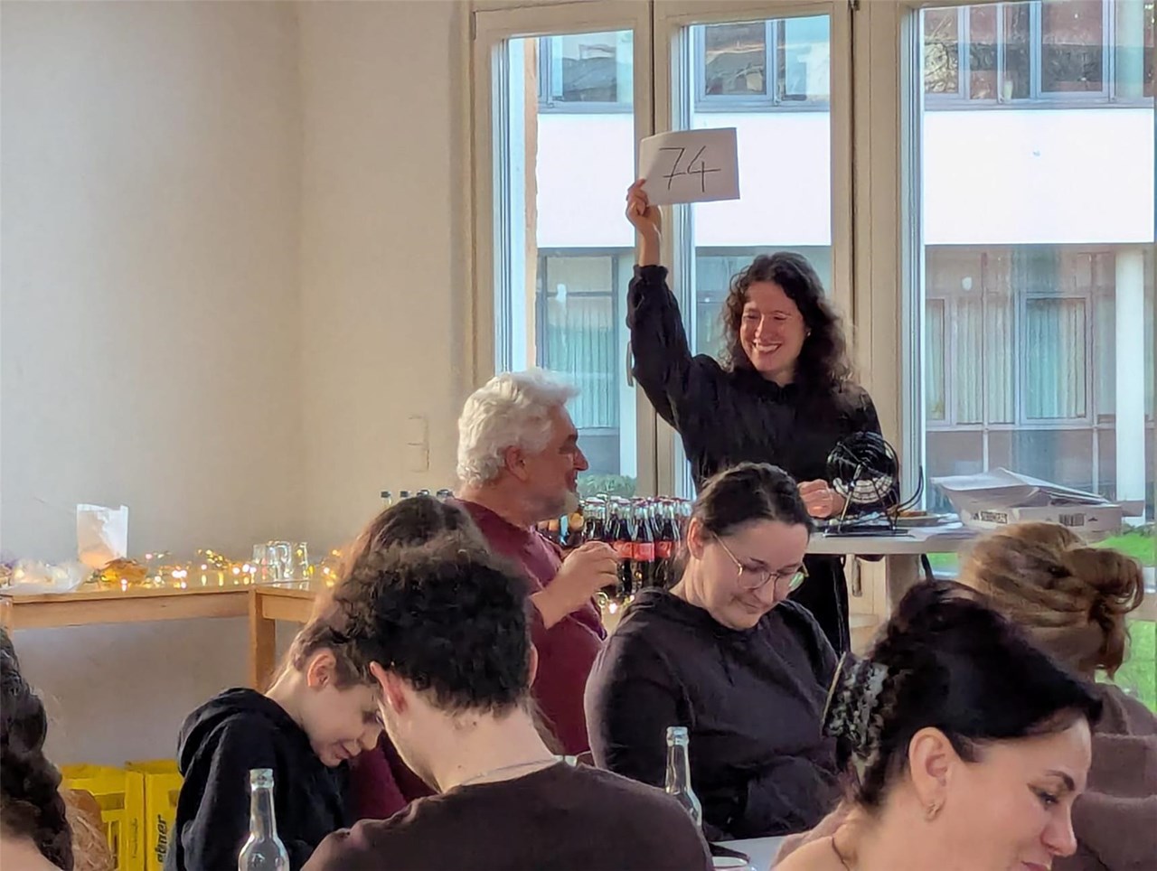 Eine Frau steht im Hintergrund und hält ein Schild mit einer Zahl hoch, andere Personen sitzen an Tischen uns spielen Bingo.