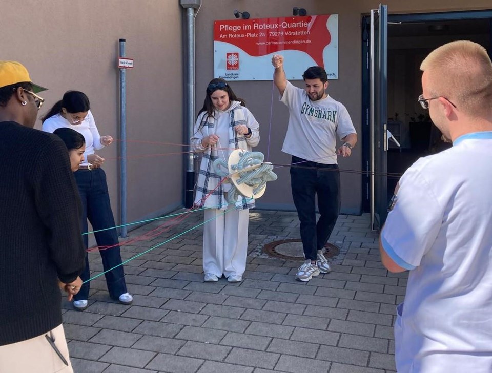 Sechs Personen, die bei einem kooperativen Teamspiel einen Gegenstand, der an Schnüren befestigt ist durch gleichmäßiges Ziehen in der Luft halten.
