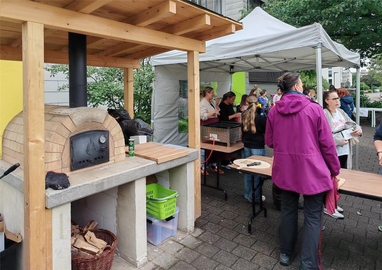 Der Pizzaofen im Vordergrund, daneben und im Hintergrund sind mehrere Personen zu sehen, die unter anderem Pizza kaufen oder verkaufen.